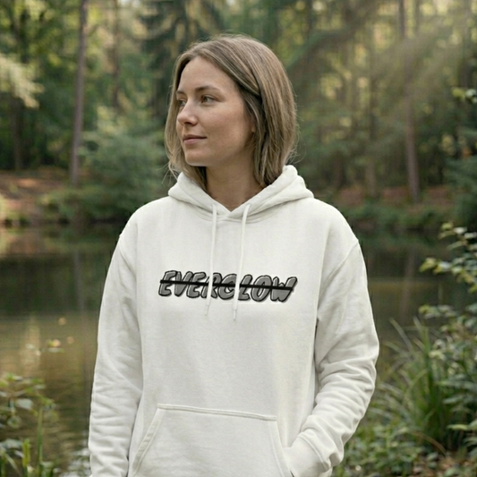 A woman in a cozy fleece Everglow: Silent Growth hoodie stands outdoors by a pond, surrounded by lush greenery and trees with sunlight streaming through the forest.