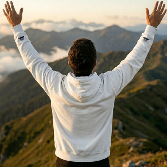 Person in white hoodie with nature graphics raising arms on mountain at sunrise