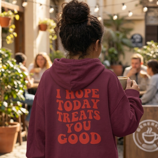 A person with curly hair in a bun, wearing the "Todays Gonna Be Good - Soft Fleece" maroon hoodie with orange affirmation text, stands outdoors holding a cup, surrounded by people and plants.