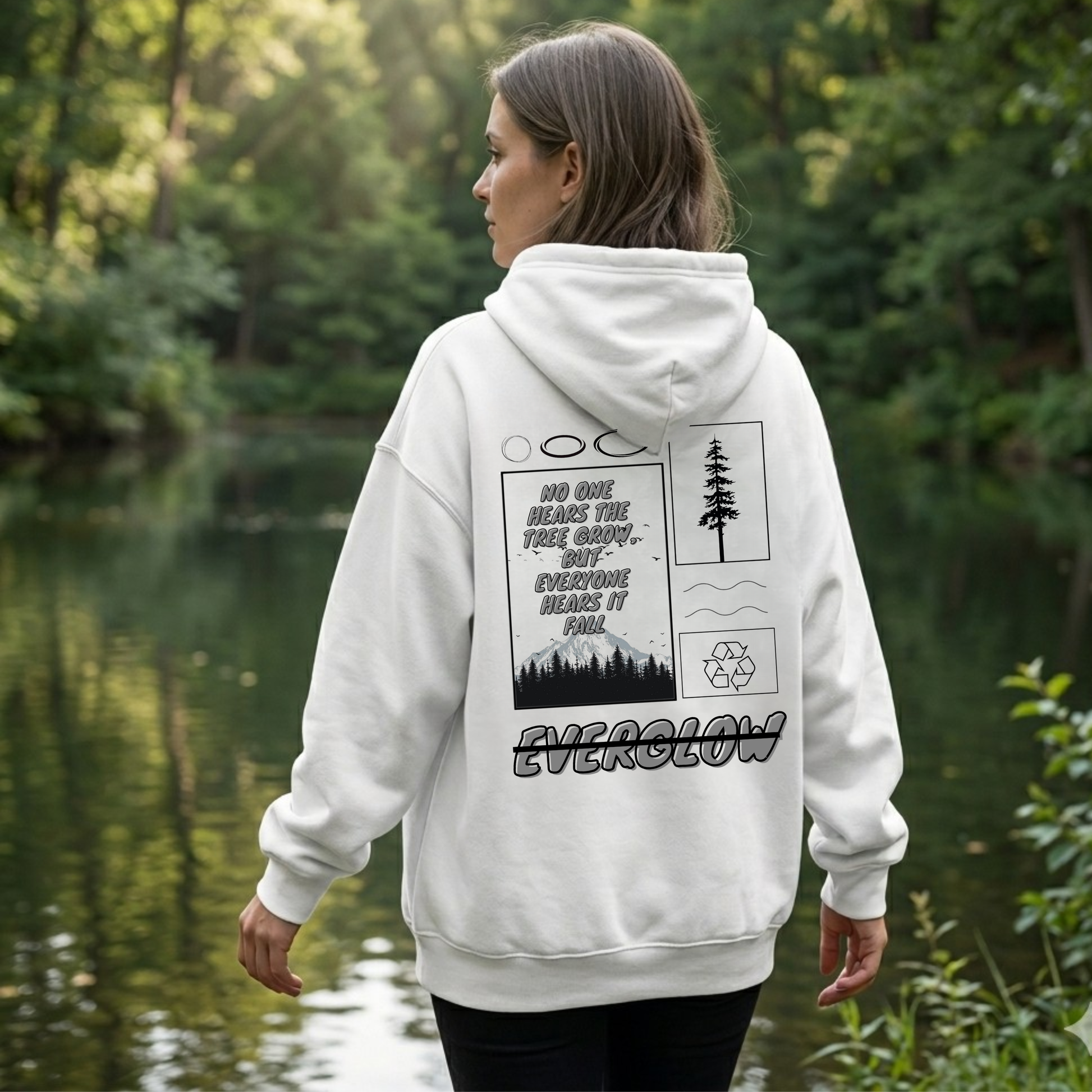 A woman in a cozy fleece hoodie stands in a green forest. The back of her Everglow: Silent Growth hoodie displays trees, mountains, a recycling symbol, and the words: No one hears the tree grow, but everyone hears it fall.