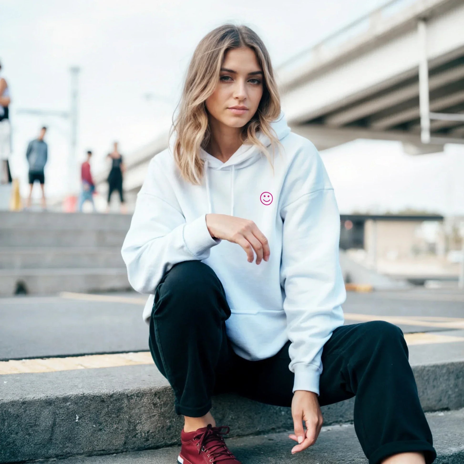 Woman in white hoodie with pink smiley graphic, black pants, and red sneakers sitting outdoors