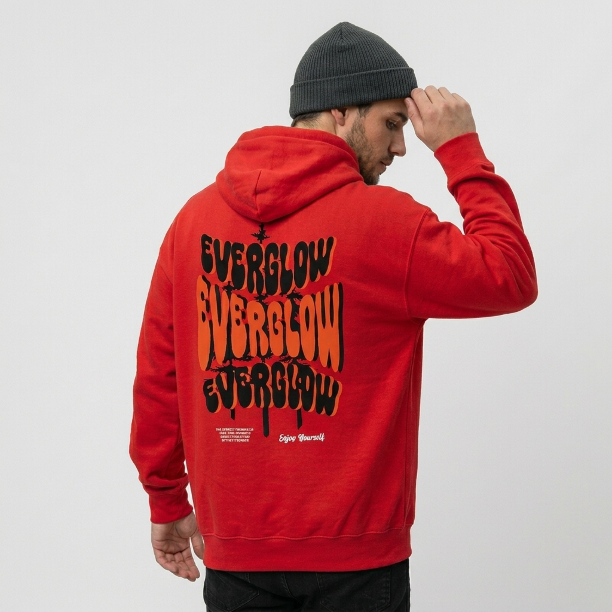A man in a gray beanie and a classic red hoodie with cozy fleece lining, featuring "Everglow: True Strength – Grow Slowly" boldly printed in wavy letters on the back, stands facing away, adjusting his hat.
