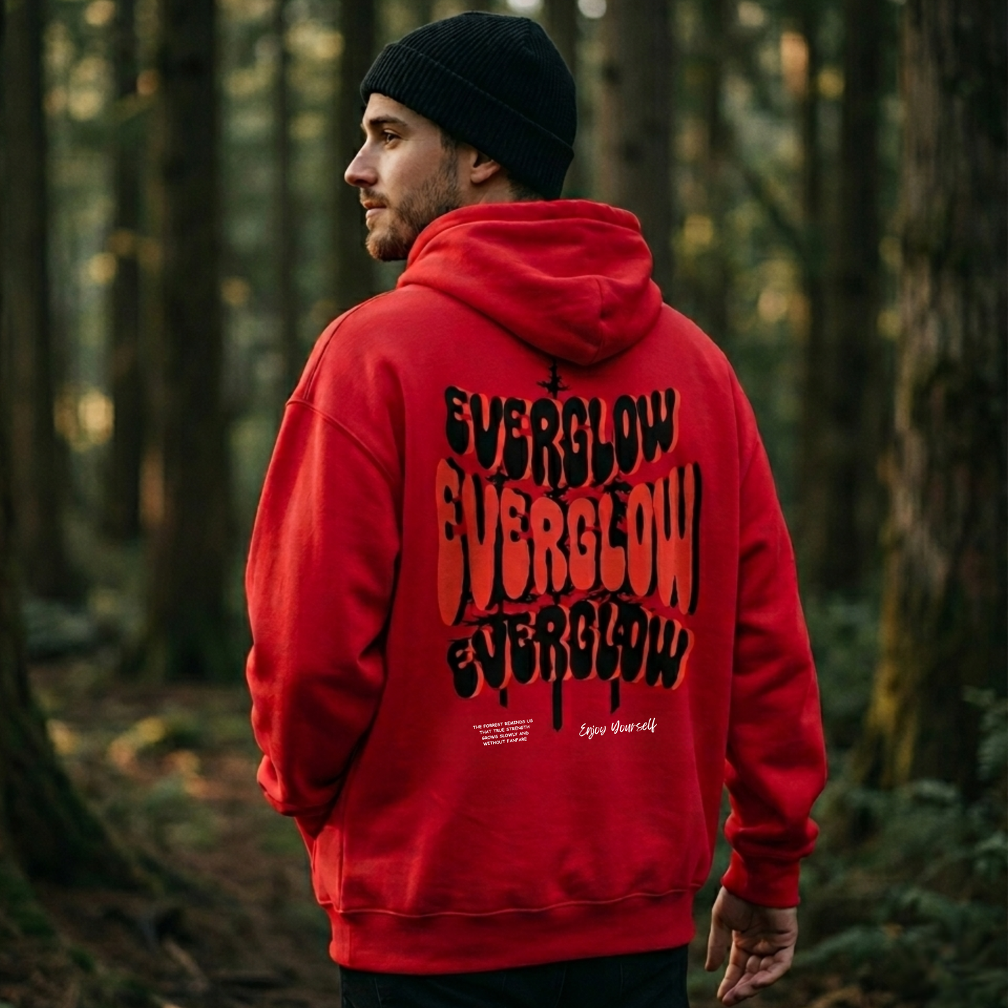 In a forest of tall trees and lush greenery, a person wearing a black beanie and the Everglow: True Strength – Grow Slowly hoodie in red with classic unisex fit stands looking to the side, EVERGLOW printed boldly on the back.