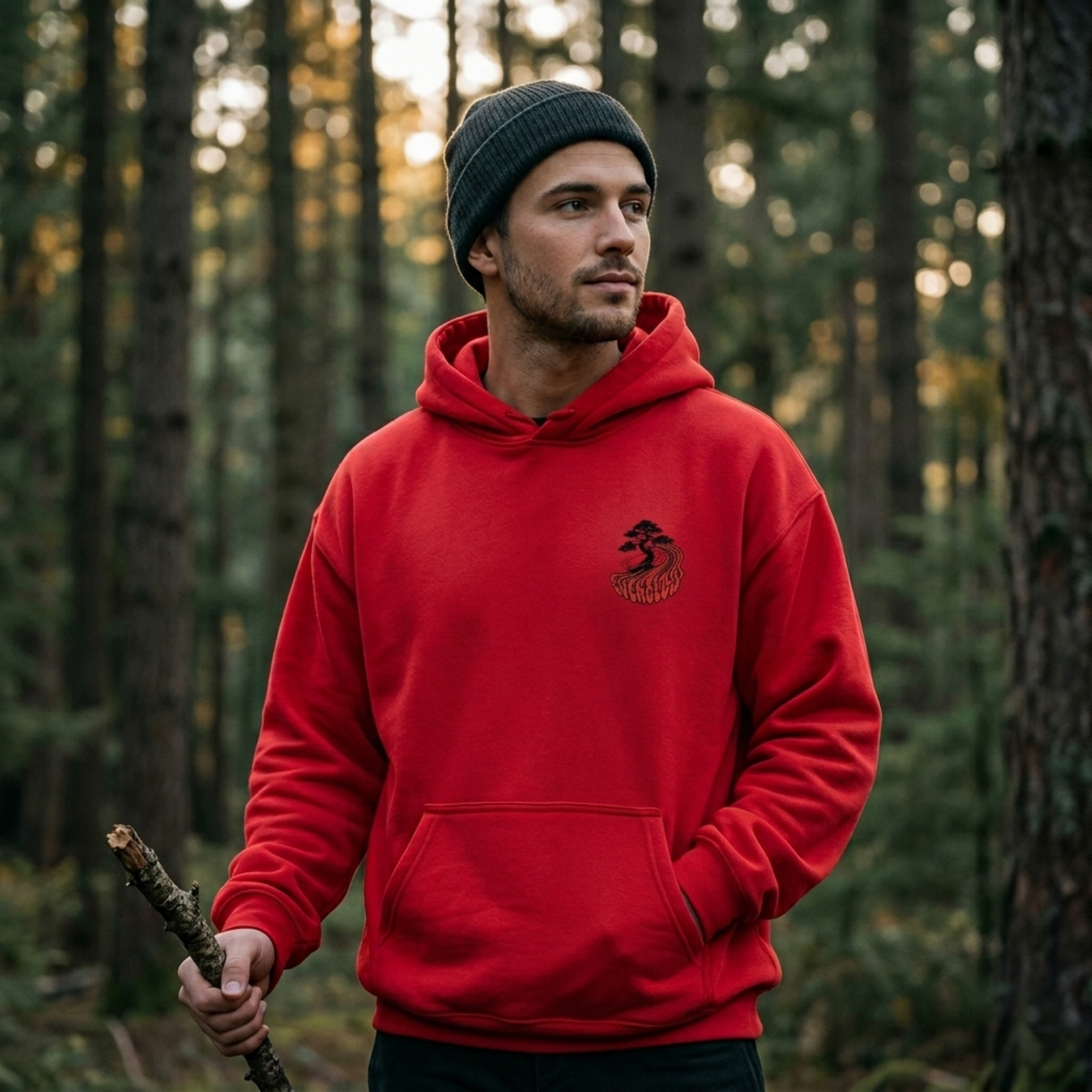 Wearing the Everglow: True Strength – Grow Slowly hoodie with cozy fleece lining and a gray beanie, a man stands in a sunlit forest, holding a stick among tall trees and lush greenery.