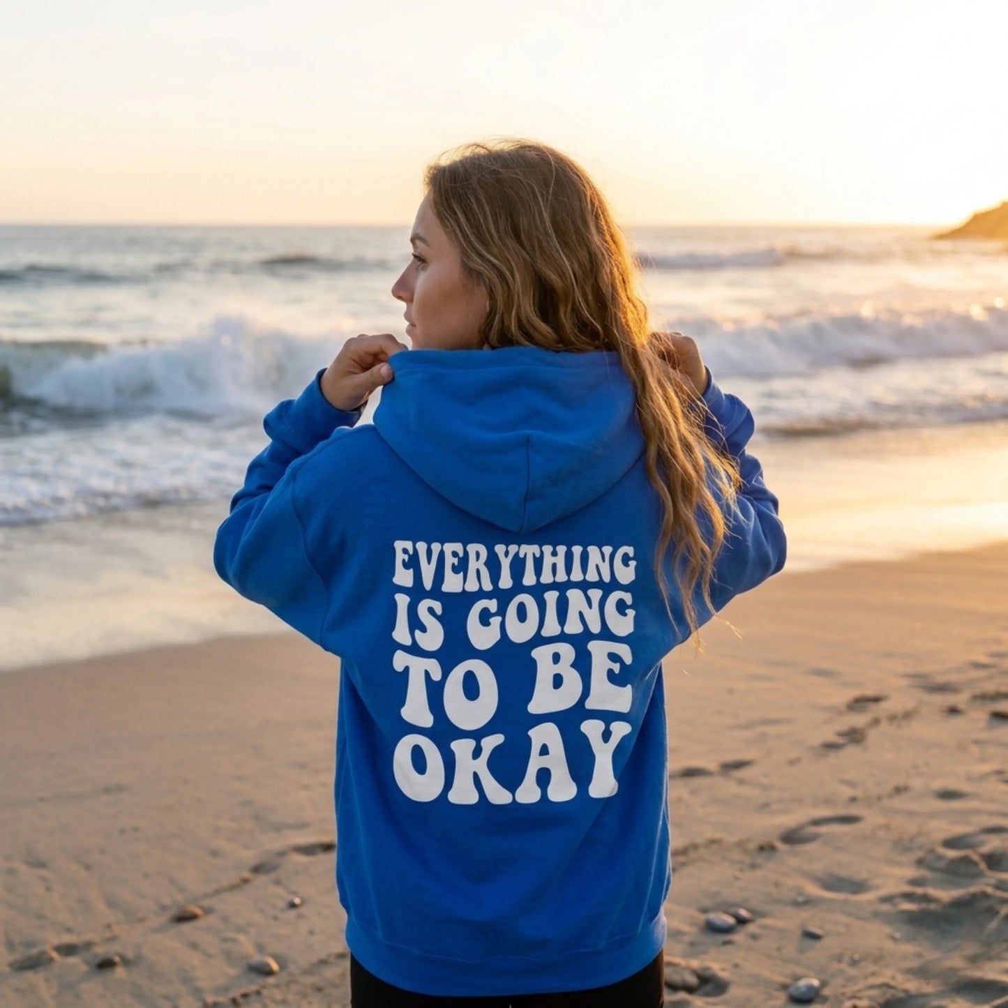 Woman in blue hoodie with 'Everything is going to be okay' text standing on beach at sunset