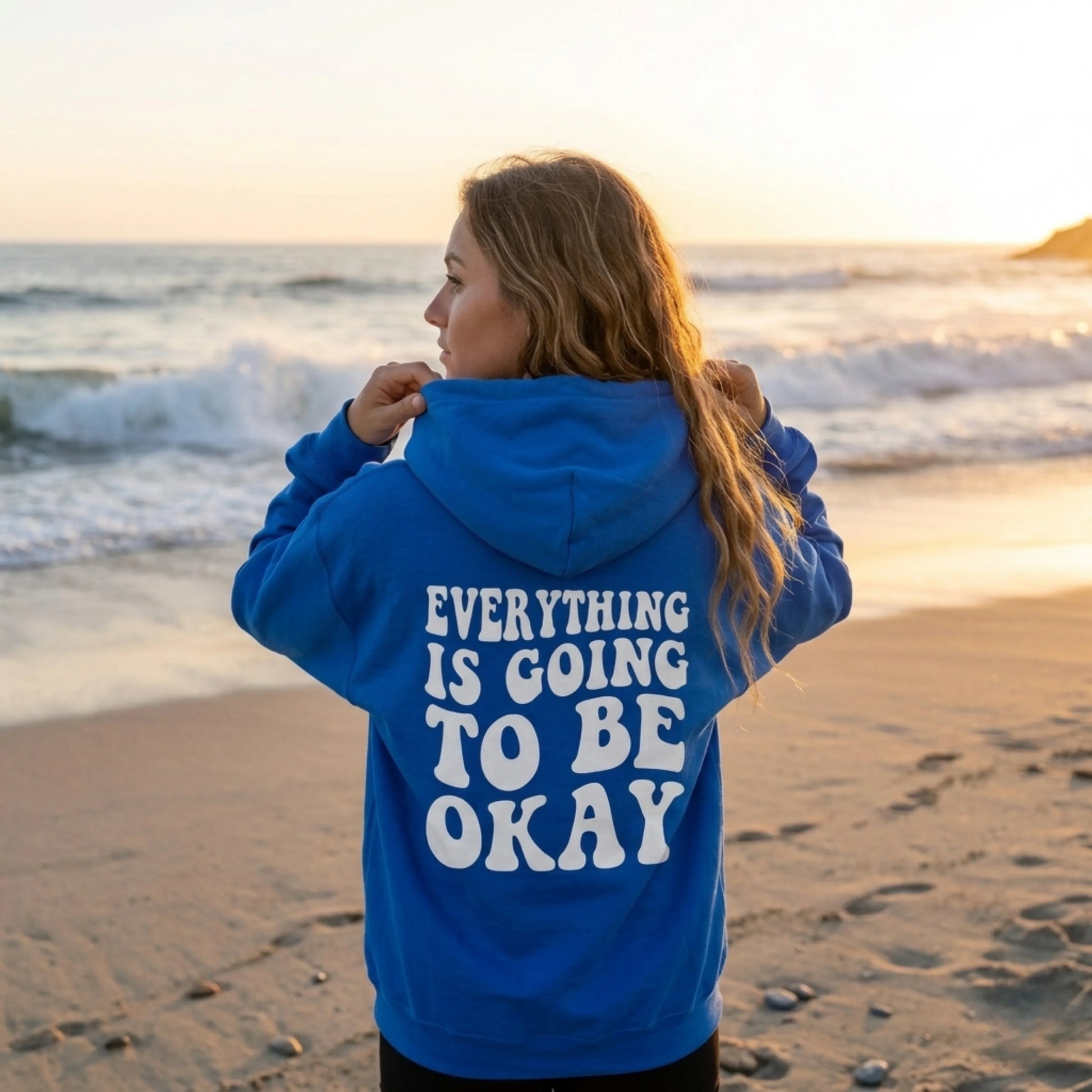 Woman in blue hoodie with 'Everything is going to be okay' text standing on beach at sunset