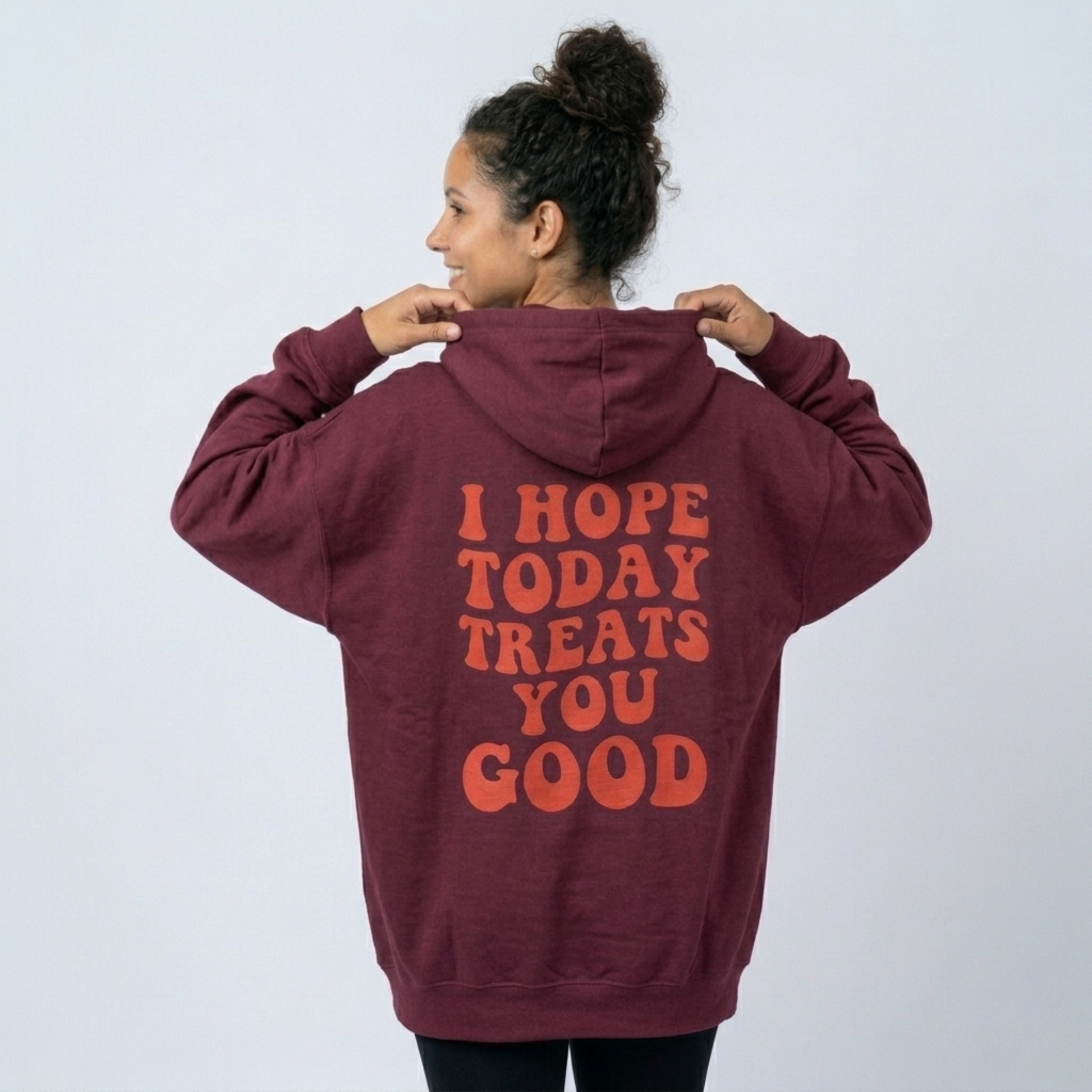 A person with curly hair in a bun smiles and looks to the side, wearing the Todays Gonna Be Good - Soft Fleece: a maroon unisex hoodie with orange "I HOPE TODAY TREATS YOU GOOD" text on the back, against a plain background.