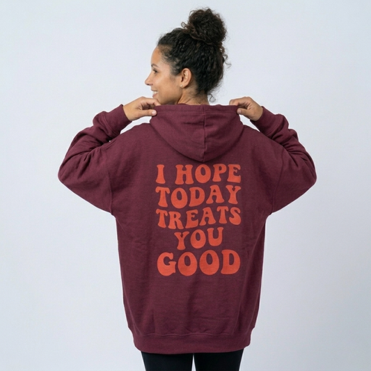 A person with curly hair in a bun smiles and looks to the side, wearing the Todays Gonna Be Good - Soft Fleece: a maroon unisex hoodie with orange "I HOPE TODAY TREATS YOU GOOD" text on the back, against a plain background.