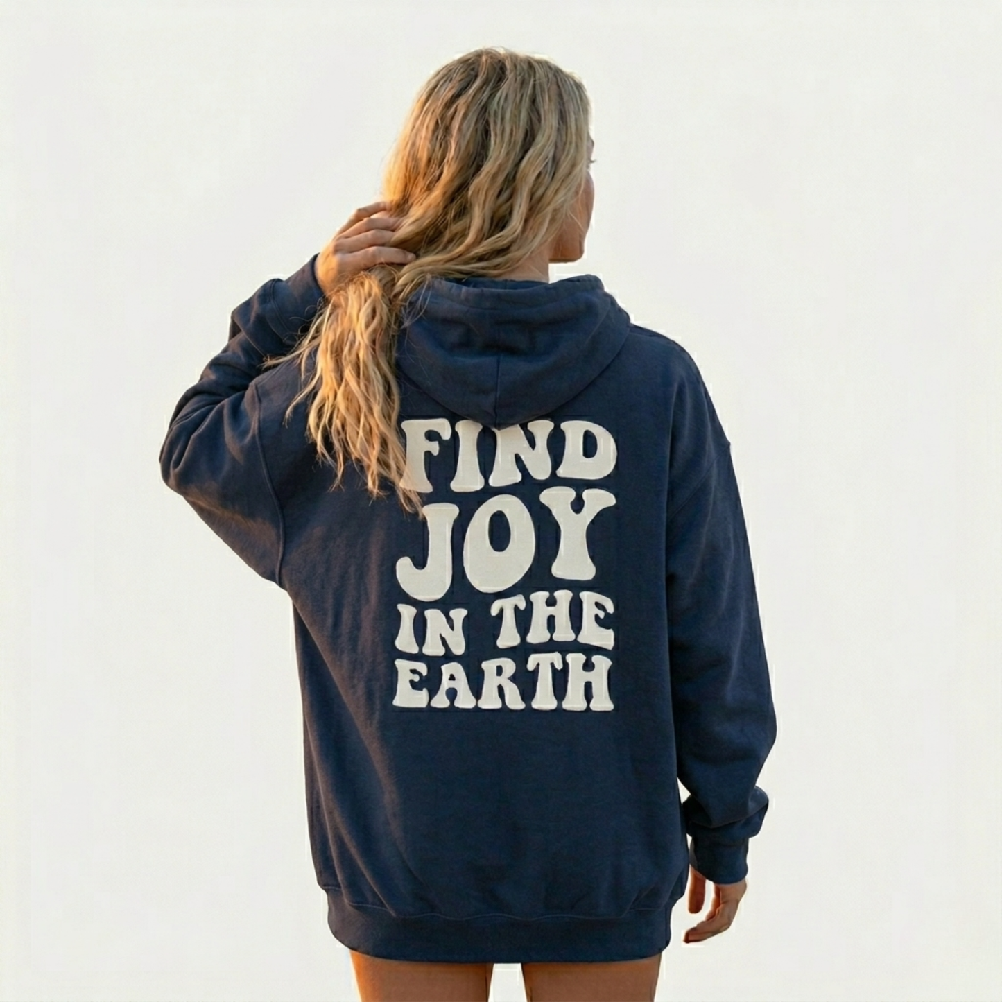 A person with long, wavy hair stands facing away, wearing a navy blue Joy In Everything - Soft Fleece hoodie with bold white lettering. The unisex relaxed fit stands out against the plain, light-colored background.