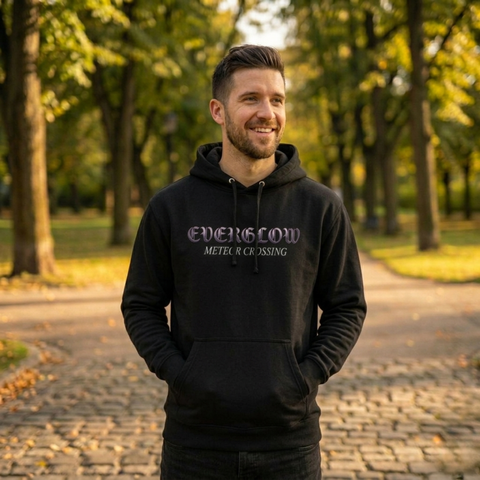A man in a black Create Memories Hoodie stands smiling on a cobblestone path in a sunlit park, surrounded by green trees.