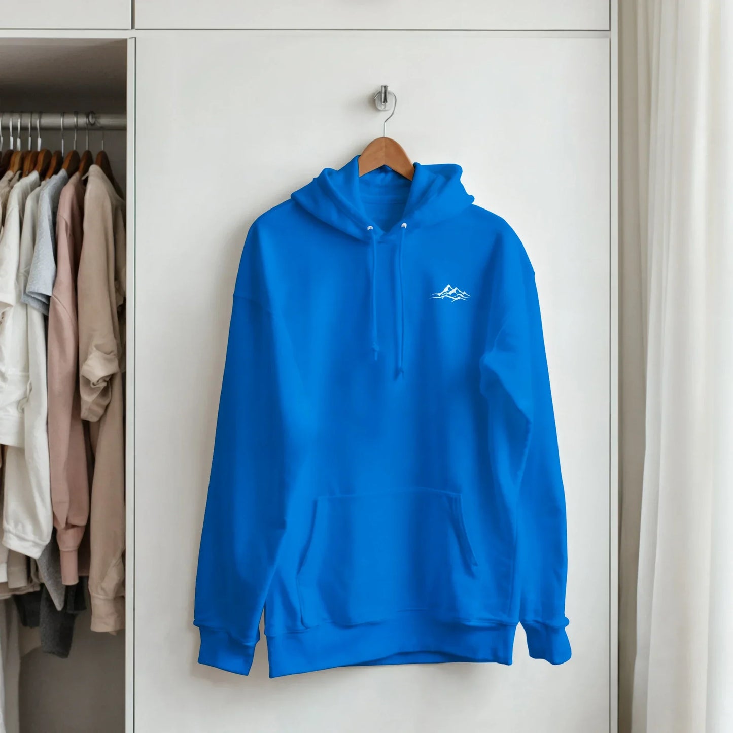 Blue unisex hoodie with small white mountain graphic, hanging in a modern closet