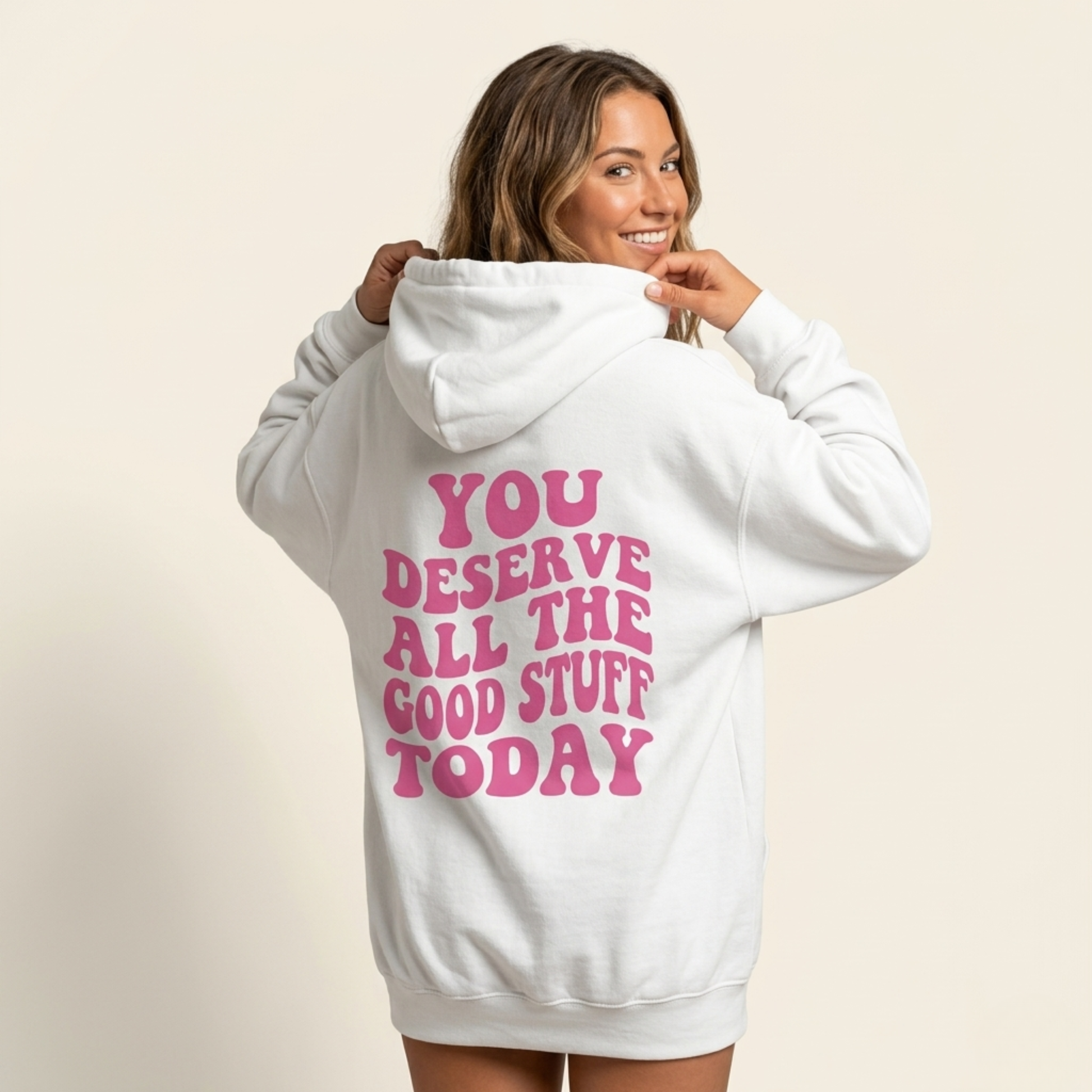 A smiling woman wears the "You Deserve All The Good Stuff Today" oversized self-love hoodie with pink text on the back, standing against a plain light background.