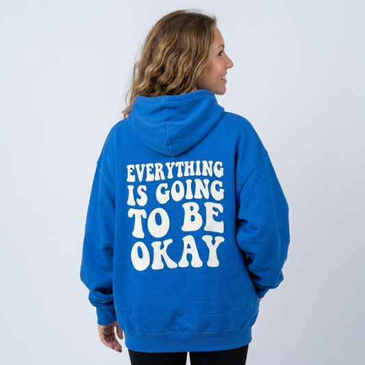 Wearing the bright blue "Everything's Gonna Be Okay - Soft Fleece" hoodie with white text on the back, a person with long, wavy hair stands against a plain light background, looking to the side and smiling.